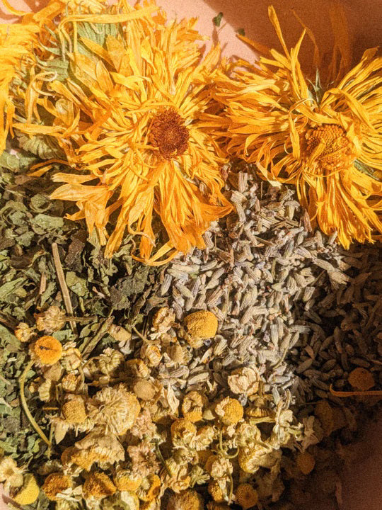 A small glass jar labeled "PINK HOUSE ORGANICS - CALM BALM" containing a light green, soothing balm. The jar is open, and the white lid is placed next to it on a neutral background. Ideal for all skin types, the balm appears smooth, indicating it hasn't been used yet.
