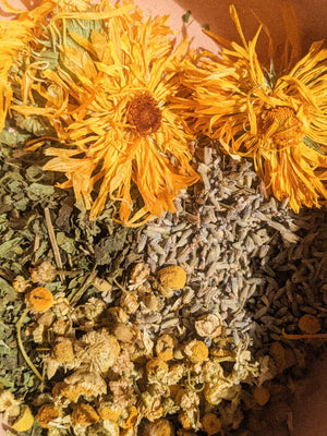 A small glass jar labeled "PINK HOUSE ORGANICS - CALM BALM" containing a light green, soothing balm. The jar is open, and the white lid is placed next to it on a neutral background. Ideal for all skin types, the balm appears smooth, indicating it hasn't been used yet.