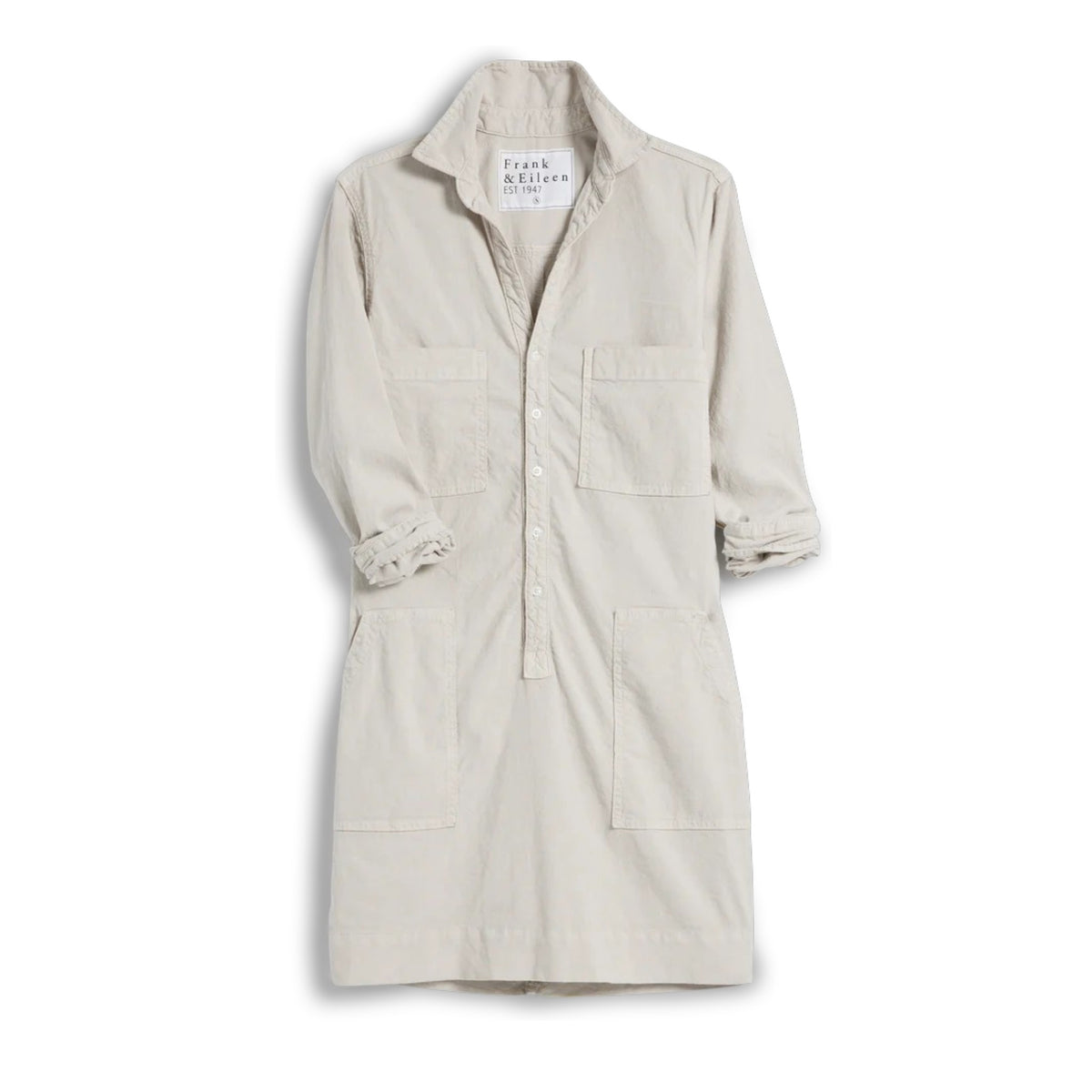 A woman stands indoors by tall windows, wearing the FRANK & EILEEN Southern Ireland Utility Dress in Performance Linen Cement. Sunlight highlights her light-colored, long-sleeve shirt dress as greenery is visible outside.
