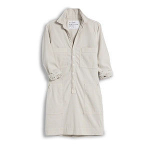 A woman stands indoors by tall windows, wearing the FRANK & EILEEN Southern Ireland Utility Dress in Performance Linen Cement. Sunlight highlights her light-colored, long-sleeve shirt dress as greenery is visible outside.