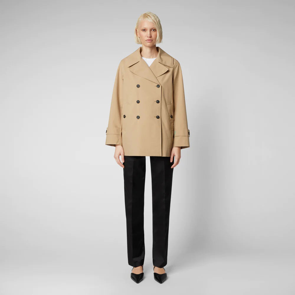 A person with short blond hair wears the SAVE THE DUCK - SOFI TRENCH JACKET by SAVE THE DUCK, a beige recycled polyester trench coat, over a white shirt with black pants and pointed shoes, standing against a light gray background.