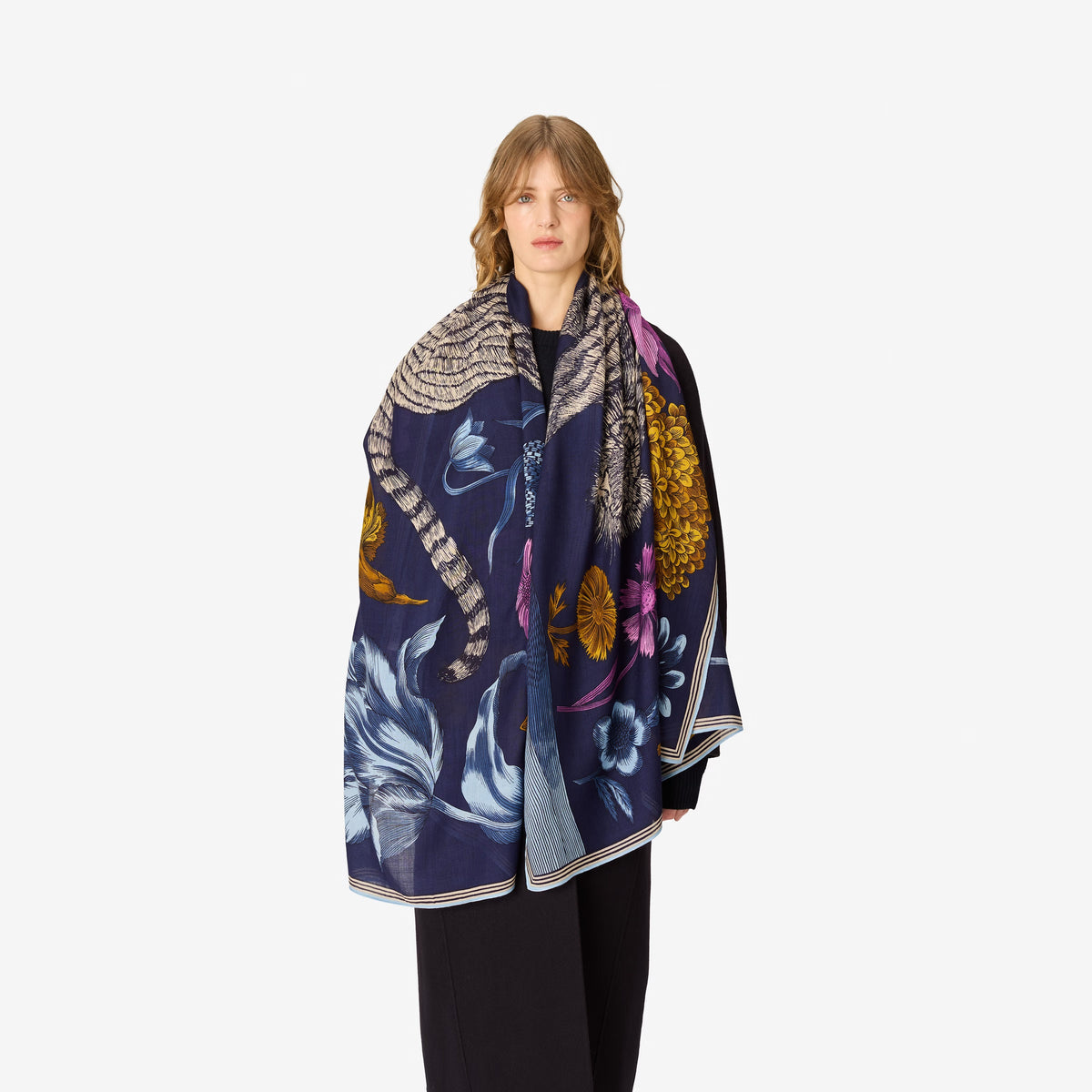 A person stands facing forward against a plain white background, wearing a long dark coat, wide pants, black shoes, and the INOUI EDITIONS - GUSTAV Wool Scarf in Navy by INOUI. An elegant wool rug made in India (80 x 190 cm) is laid beneath their feet.