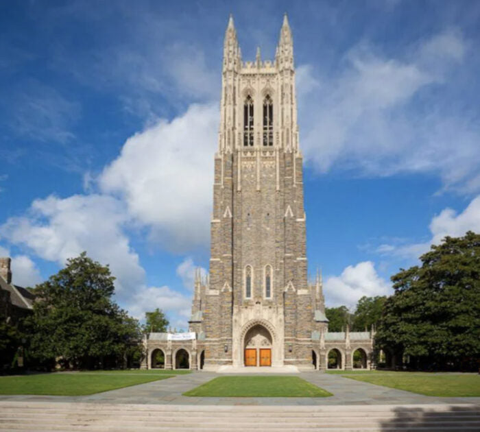 A tall stone Gothic-style chapel with a central tower stands amid green lawns and trees under a partly cloudy blue sky. Wide steps lead up to wooden doors at the building’s entrance.