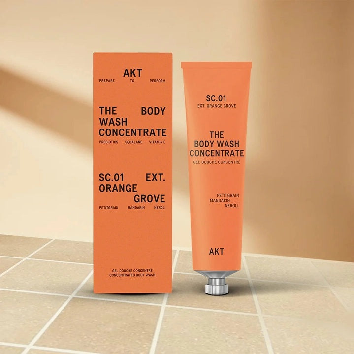 An orange tube of AKT LONDON’s BODY WASH CONCENTRATE ORANGE GROVE is squeezed onto a hand. Beside it, on an orange background, text reads: “SKIN-BALANCING,” “MOISTURE-LOCKING,” “NOURISHING.”.