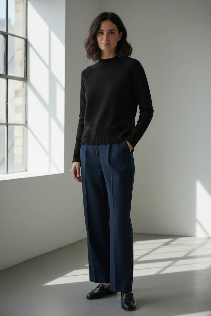 A person with long light brown hair is wearing the KINROSS CASHMERE Hi Low Rib Trim Funnel Cashmere Sweater in Black by KINROSS, paired with dark pants. They stand against a light background with one hand in their pocket, looking at the camera and smiling slightly.