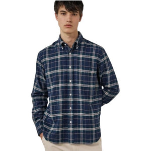 A young person with dark hair, wearing the HARTFORD - PITT CHECKED FLANNEL SHIRT IN BLUE AND GREEN by HARTFORD, looks at the camera while adjusting the shirt collar with one hand against a plain light background.