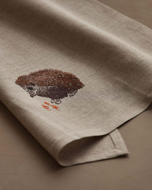A HEDGEHOG WITH MUSHROOMS LINEN TEA TOWEL by CORAL & TUSK hangs from the handle of a white stove in a kitchen.