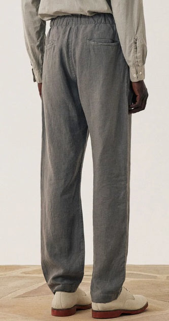 A man stands against a plain background wearing a light gray long-sleeve henley shirt, HARTFORD - LINEN JOGGY PANTS IN ARMY, and light-colored shoes, with his hands in his pockets.