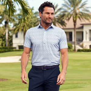 A man in the COLLARS AND CO Featherweight Flex Polo in Coastal Blue Floral with a dress collar and dark pants stands outdoors by a large pillar, with green grass, gardens, and a building in the background.