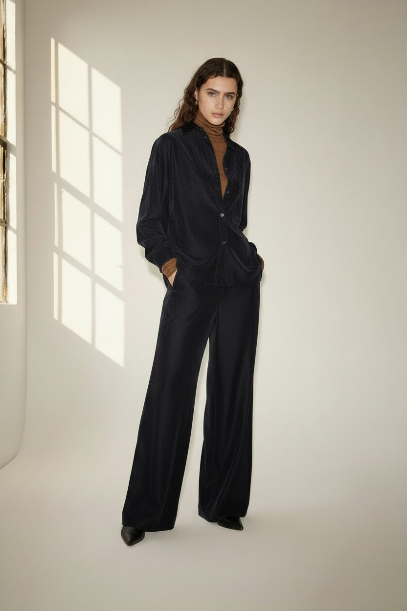 A person with long, wavy brown hair wears a loose-fitting, dark velvet suit over a brown top and stands in a softly-lit room with a large window casting light and shadows on the wall. The look is completed with NUDE’s Wide Leg Trouser in Blue, adding an effortless elegance to the ensemble.
