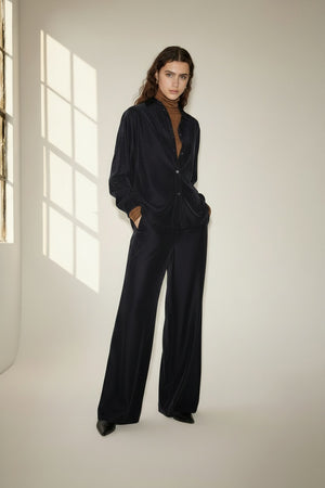 A person with long, wavy brown hair wears a loose-fitting, dark velvet suit over a brown top and stands in a softly-lit room with a large window casting light and shadows on the wall. The look is completed with NUDE’s Wide Leg Trouser in Blue, adding an effortless elegance to the ensemble.