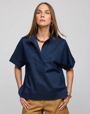 A woman with long brown hair is seen wearing the HINSON WU Cara Elbow Sleeve Shirt in Stretch Polished Cotton Navy—a loose-fitting, oversized short-sleeve shirt—paired with tan pants and gold jewelry, standing against a plain light gray background.