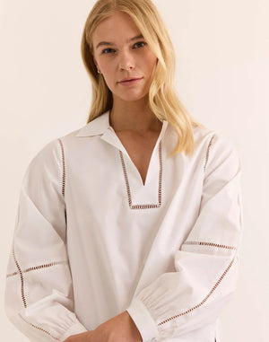 A woman with straight blonde hair wears the HINSON WU Laura Long Sleeve Lace Trim Top in White, paired with black pants, standing against a plain light background.