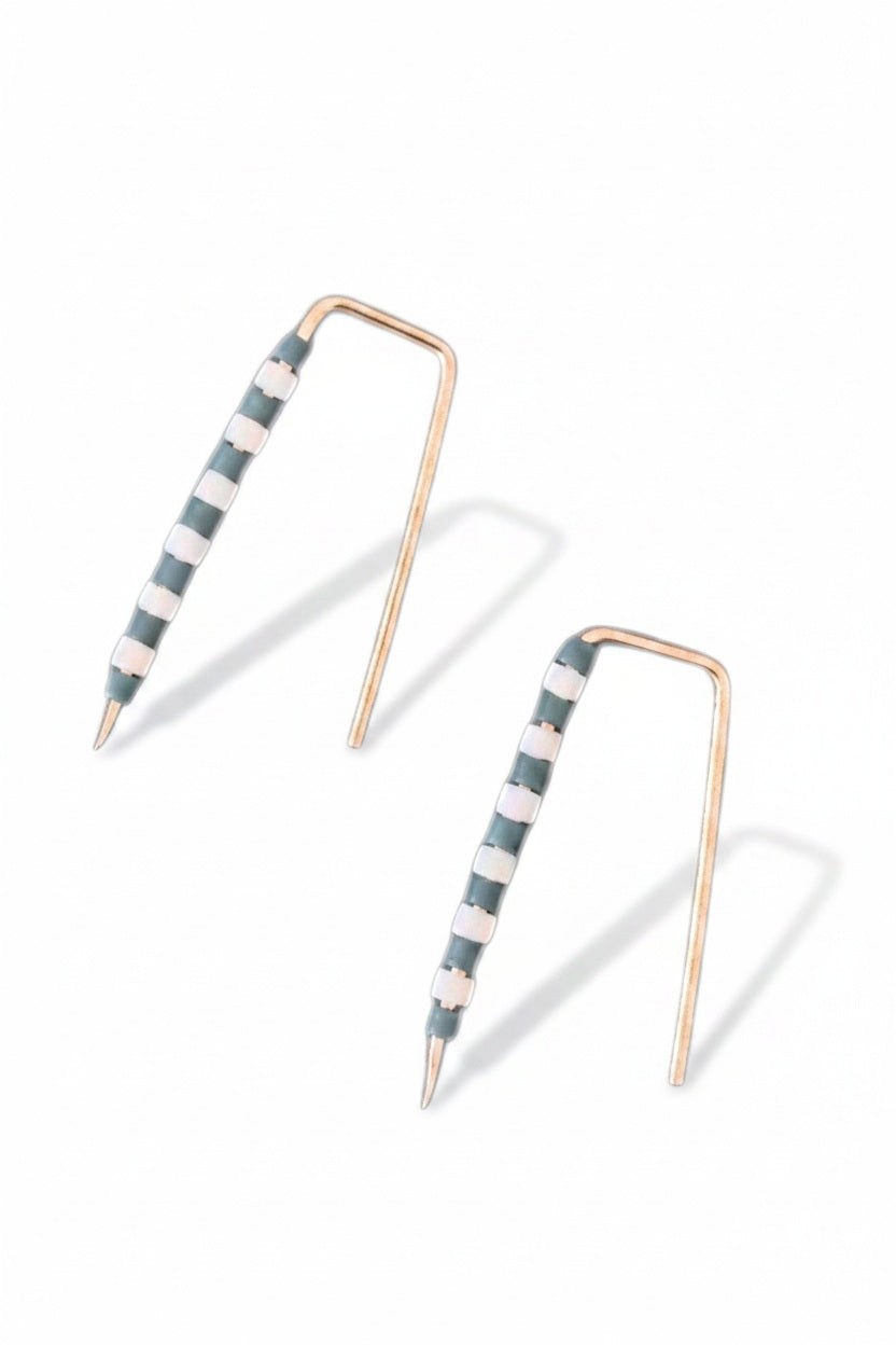 The ALICE RISE - LITTLE QUILL EARRINGS IN SLATE BLUE STRIPE feature unique 14k gold filled posts adorned with alternating white and green beads, arranged parallel on a textured light gray surface.