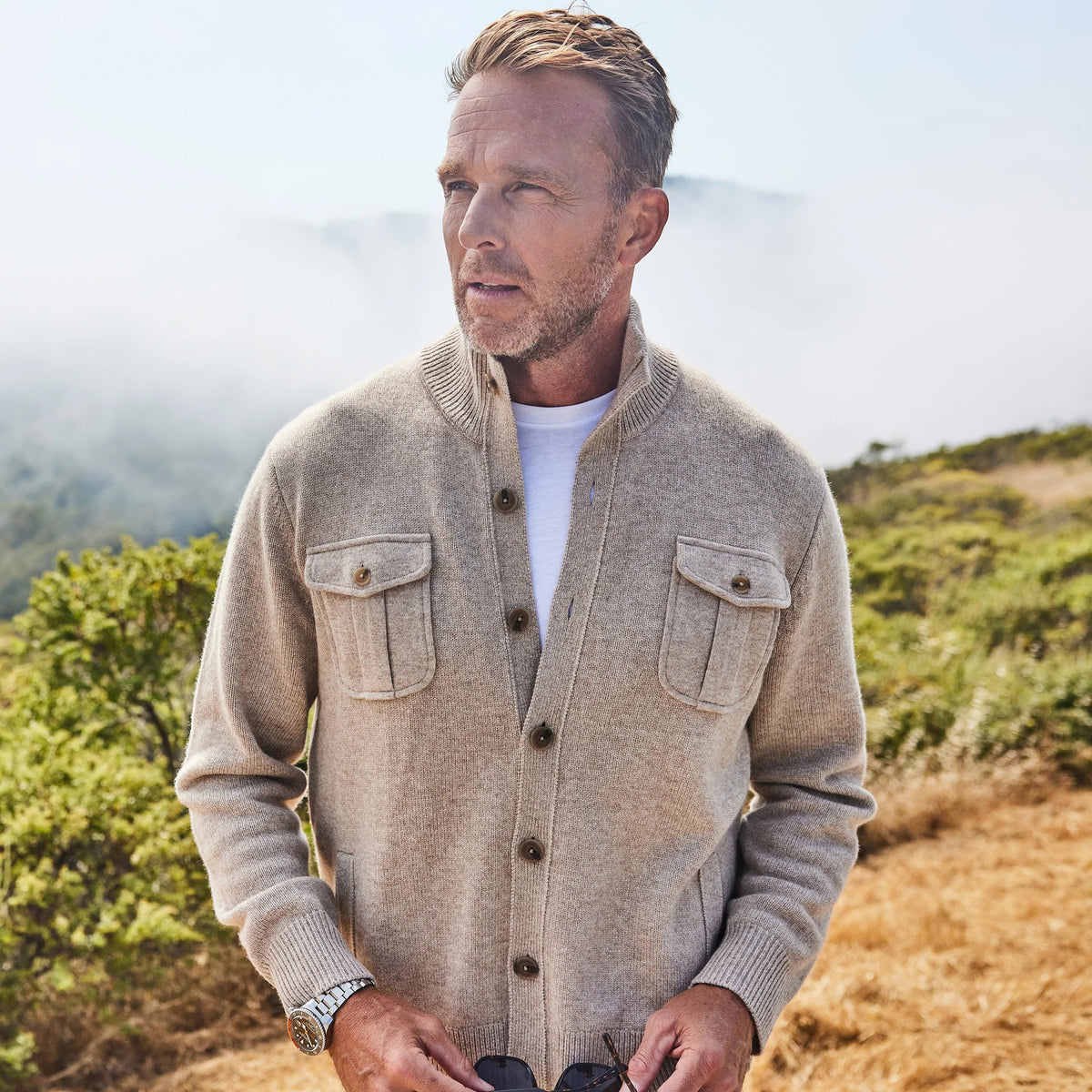 A man with short hair and a trimmed beard stands outdoors, wearing the COLLARS AND CO - NAPOLI CARDIGAN IN WOOL AND CASHMERE CAMEL over a white shirt, looking seriously at the camera with misty hills and green bushes in the background.