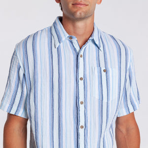 A man wearing the CRIQUET - THE BEACHCOMBER SHIRT IN BLUE by CRIQUET, paired with light beige shorts, stands against a plain white background, looking to his left with one hand in his pocket.