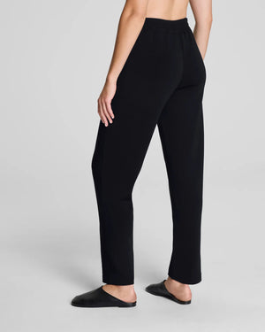 A person models the SPANX - AIRessentials Brushed Slim Straight Pant with an elastic waistband and buttons, paired with black slip-on shoes against a plain light background.