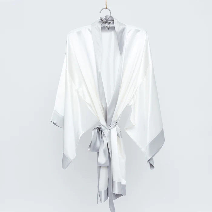 The BRANCHE GEISHA Robe Silk in pink satin, adorned with white trim, hangs gracefully on a matching silk hanger. This elegant robe by BRANCHE features long, wide sleeves and is accentuated with a white sash tied in a bow around the waist, complete with mitered corners for an exquisite finish.