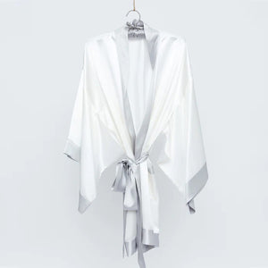 The BRANCHE GEISHA Robe Silk in pink satin, adorned with white trim, hangs gracefully on a matching silk hanger. This elegant robe by BRANCHE features long, wide sleeves and is accentuated with a white sash tied in a bow around the waist, complete with mitered corners for an exquisite finish.