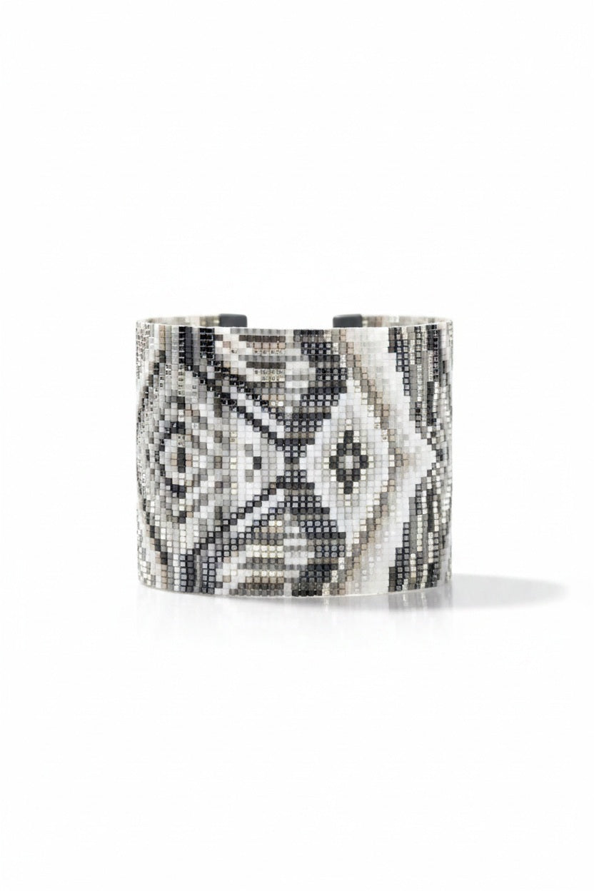 The JULIE ROFMAN - HORIZON CUFF from JULIE ROFMAN JEWELRY is a handwoven glass beaded delica bracelet that showcases an intricate geometric pattern in black, white, and gray. The design includes diamond shapes and symmetrical lines for a mosaic-like appearance, finished with an adjustable buckle clasp to ensure a perfect fit.
