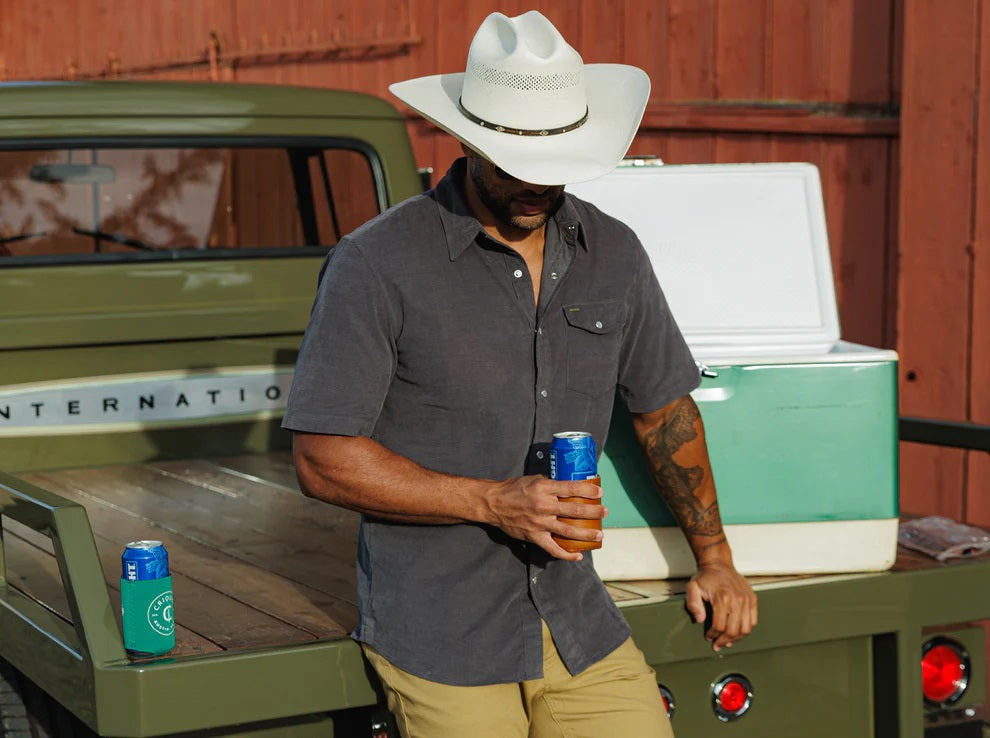 A smiling man in a brown hat and the CRIQUET Short Sleeve Corduroy Pearl Snap Shirt in Iron Horse holds a canned drink in his right hand, standing against a white background.