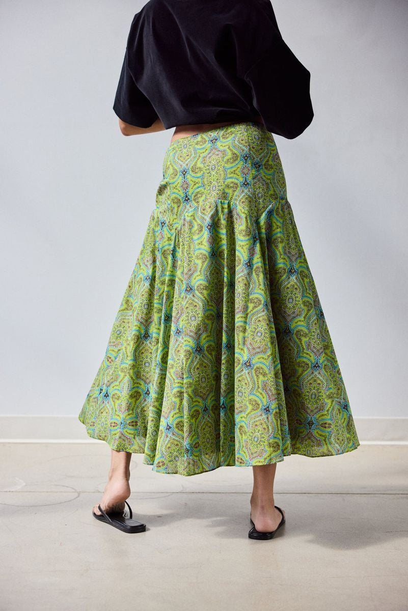A woman stands indoors in a loose black top and the HOPE FOR FLOWERS - CIRCLE SKIRT IN GREEN FOULARD, made from soft organic cotton lawn. She holds out one side of her GOTS certified skirt by HOPE FOR FLOWERS, paired with black sandals.