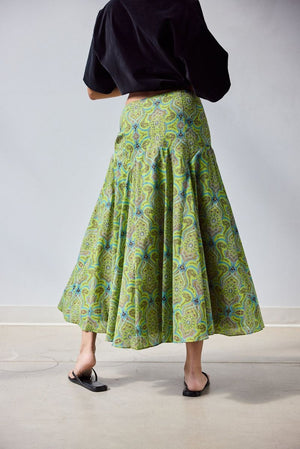 A woman stands indoors in a loose black top and the HOPE FOR FLOWERS - CIRCLE SKIRT IN GREEN FOULARD, made from soft organic cotton lawn. She holds out one side of her GOTS certified skirt by HOPE FOR FLOWERS, paired with black sandals.