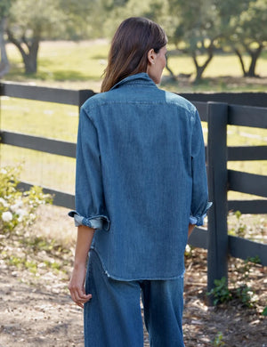 A person with long brown hair stands outdoors, facing away, next to a black wooden fence with trees and grass in the background, wearing the FRANK & EILEEN EILEEN Relaxed Button Up Shirt in Famous Denim Distressed Indigo Wash.