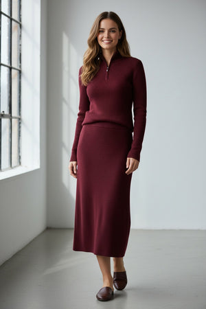 A person with long, light brown hair stands against a plain white background wearing the KINROSS cashmere Rib Zip Mock Sweater in Cordovan—a burgundy ribbed knit pullover with a zipped mock neck from KINROSS—paired with a matching skirt.
