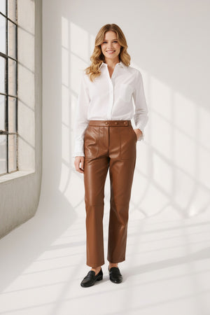A person wearing high-waisted brown leather pants, a white long-sleeve shirt, and brown heeled ankle boots, standing against a plain light background. The person's face is not visible.