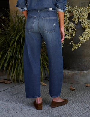 A close-up of a person wearing the FRANK & EILEEN Brennan Everyday Barrel Jean in California Vintage Denim 1993 Wash, paired with a loose blue shirt and one arm resting by their side.