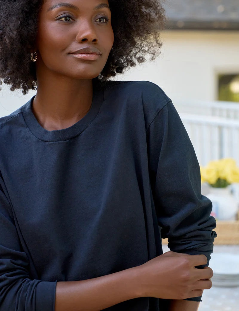 A woman with naturally curly hair, wearing the FRANK & EILEEN QUINN Long Sleeve Crewneck Tee in British Royal Navy and relaxed blue jeans, stands by an outdoor table with plates, drinks, and flowers, smiling softly. Made in California.