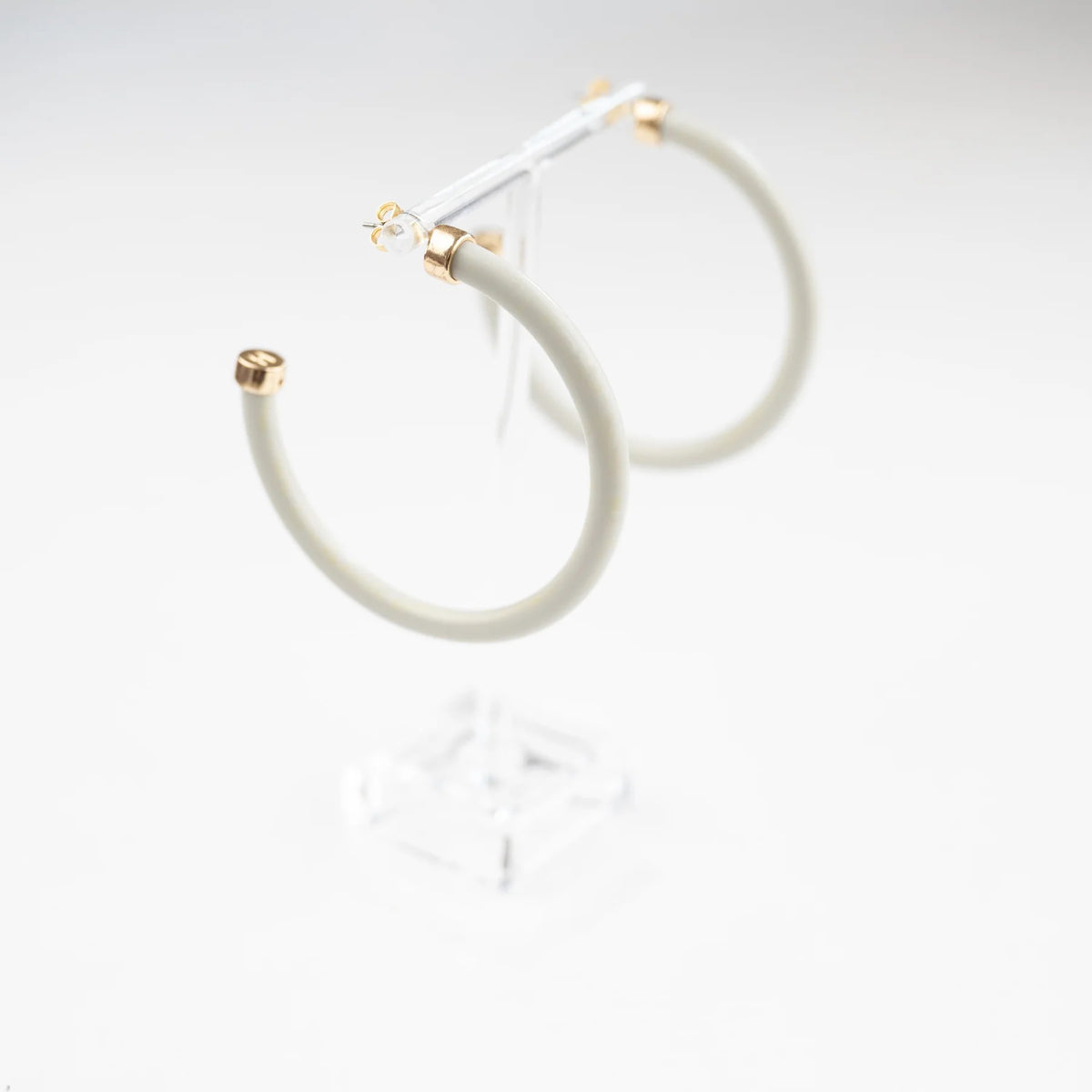 Gold and beige BONE HOOP EARRINGS by HOO HOOPS, these statement earrings are elegantly displayed on a transparent acrylic stand against a white background.