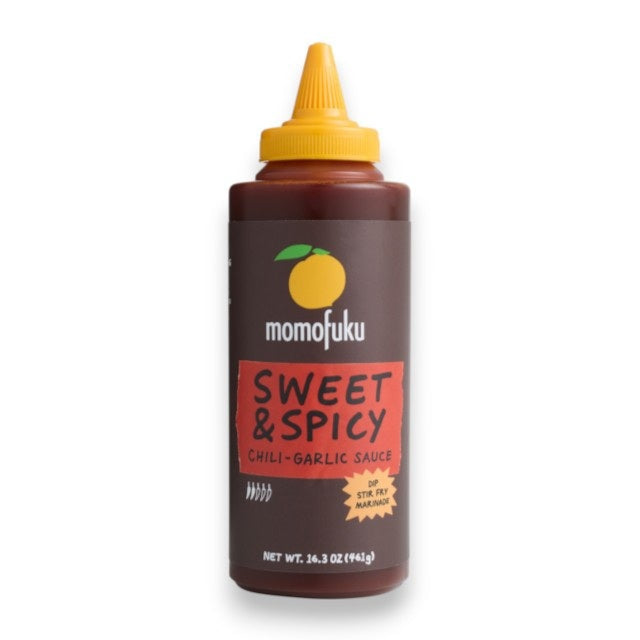 Two bottles of MOMOFUKU - SWEET & SPICY KOREAN BBQ SAUCE from MOMOFUKU, each with a yellow cap and a label featuring a yellow plum graphic, are displayed against a red and beige background—perfect for use as a vegan marinade or for adding bold, gochujang-inspired flavor to your favorite dishes.