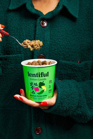 A container of LENTIFUL - MEXICAN GREEN CHILE INSTANT LENTILS is surrounded by fresh ingredients including green bell peppers, jalapeños, limes, cilantro, garlic cloves, onion slices, and green peas—all arranged on a vibrant green background.