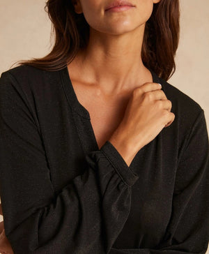 A person stands in a lavishly decorated room, wearing the shimmering long MOMONI - AMEDE DRESS IN LUREX JERSEY, made in Italy by MOMONI, paired with a black wide-brimmed hat adorned with a colorful band. Their hands are in their pockets, and there's a large mirror reflecting part of the ornate wall design.