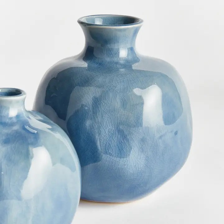 Two round, glossy blue ceramic NAPA HOME AND GARDEN - BLUE NARINE BUD VASES (Large) with narrow openings and crackle glaze patterns, positioned closely together on a plain white background.