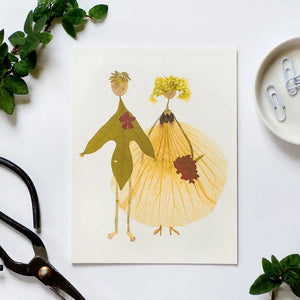 The PETAL PEOPLE PRESS - BETROTHED CARD by PETAL PEOPLE PRESS features two figures in formal wear crafted from real leaves and petals. Shown on a white surface with clippers, greenery, and paperclips nearby.