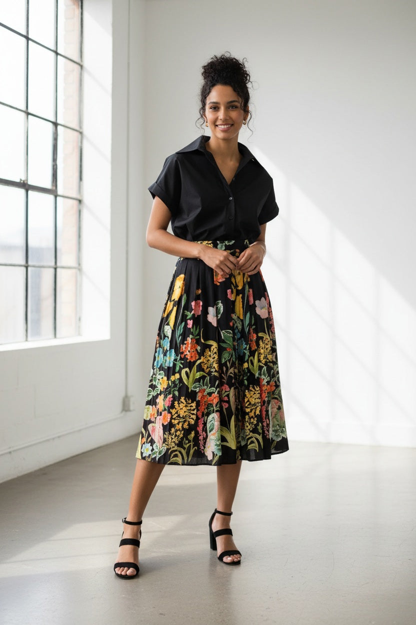 A woman stands against a white background wearing a black short-sleeve shirt paired with the LAVI - IRIS Botanical Skirt in Black Floral from LAVI, featuring colorful floral patterns, and completes the look with black high-heeled sandals.