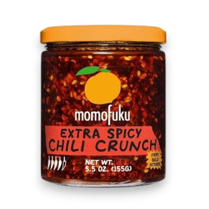 A glass jar of MOMOFUKU - EXTRA SPICY CHILI CRUNCH from MOMOFUKU has a silver lid and a label with a yellow peach, bold red and white text, and shows the spicy chili oil with Mexican chilis and red pepper flakes inside.