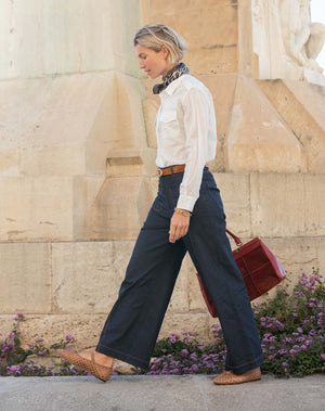 A woman with curly hair stands smiling in a bright room, wearing the MERSEA Andie Woven Pant in Rinse—high-waisted, wide-leg trousers with front pockets—paired with a white button-up shirt tucked in, a brown belt, and brown woven flats. One hand rests in her pocket.