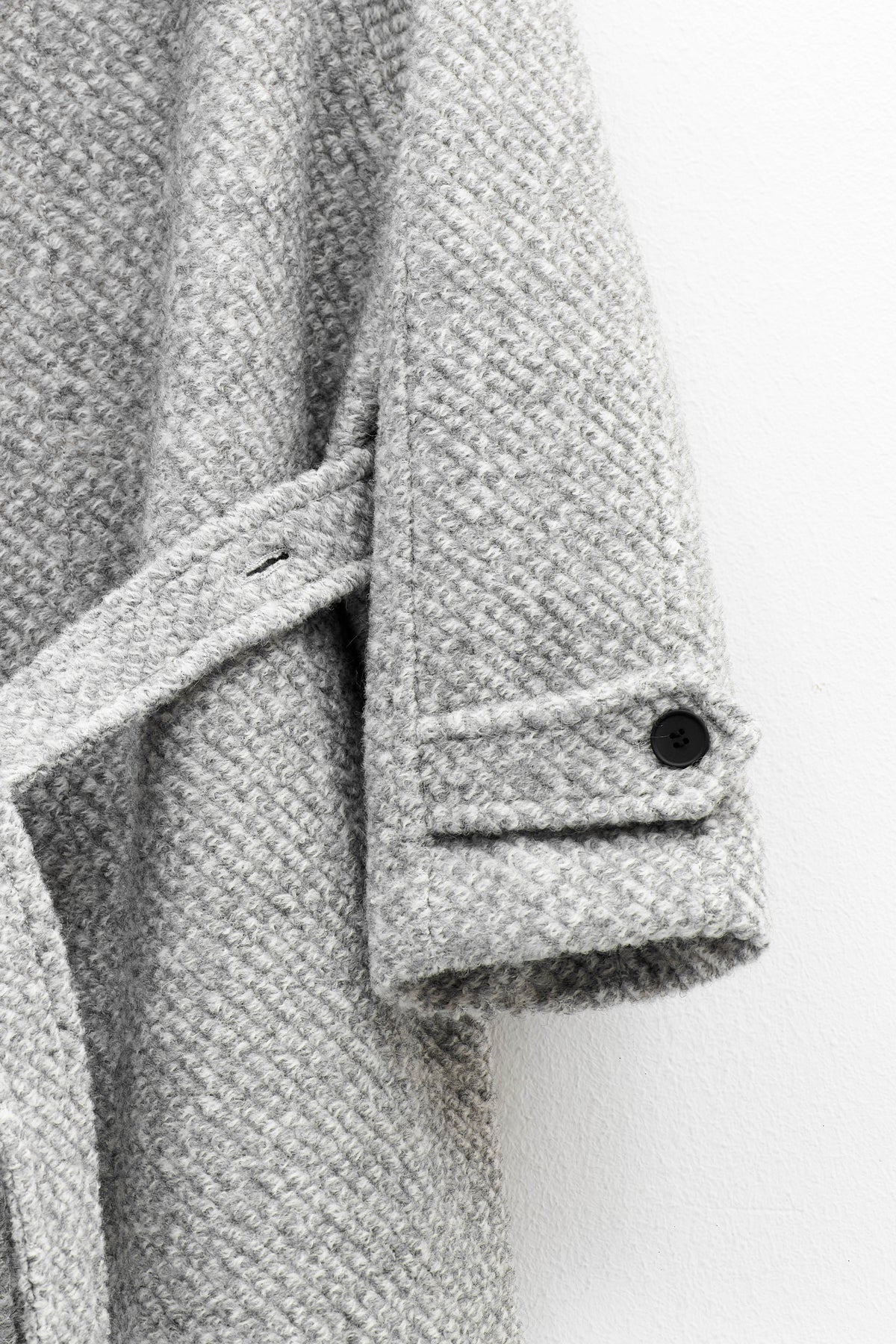 Close-up of the ELEH - THE AURELIA COAT in light gray wool blend, showing a fabric belt and black cuff button, hanging against a white wall.
