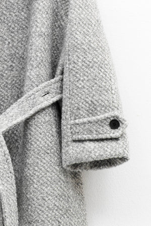 Close-up of the ELEH - THE AURELIA COAT in light gray wool blend, showing a fabric belt and black cuff button, hanging against a white wall.