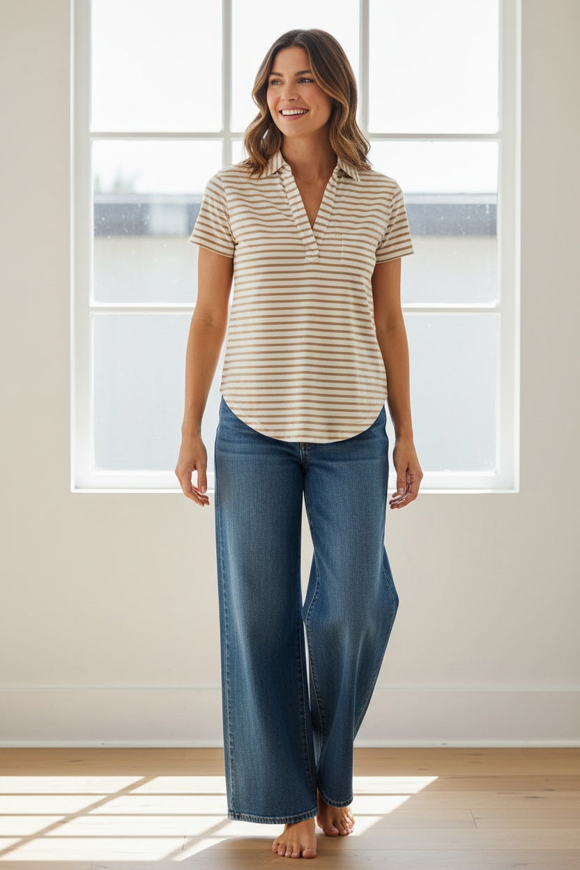 A woman with blonde hair stands outdoors, looking at the camera, wearing the FRANK &amp; EILEEN Charlotte Perfect Polo in Heritage Jersey Wintage White and Camel Stripe. Greenery and white flowers are softly blurred in the background.