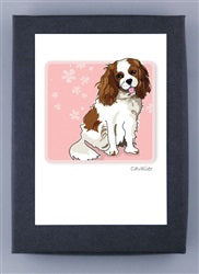 The PAPER RUSSELLS CAVALIER KING CHARLES SPANIEL NOTE CARDS feature a brown and white dog with long, fluffy ears and a playful expression against a pink background with floral designs on recycled paper, bordered in black.