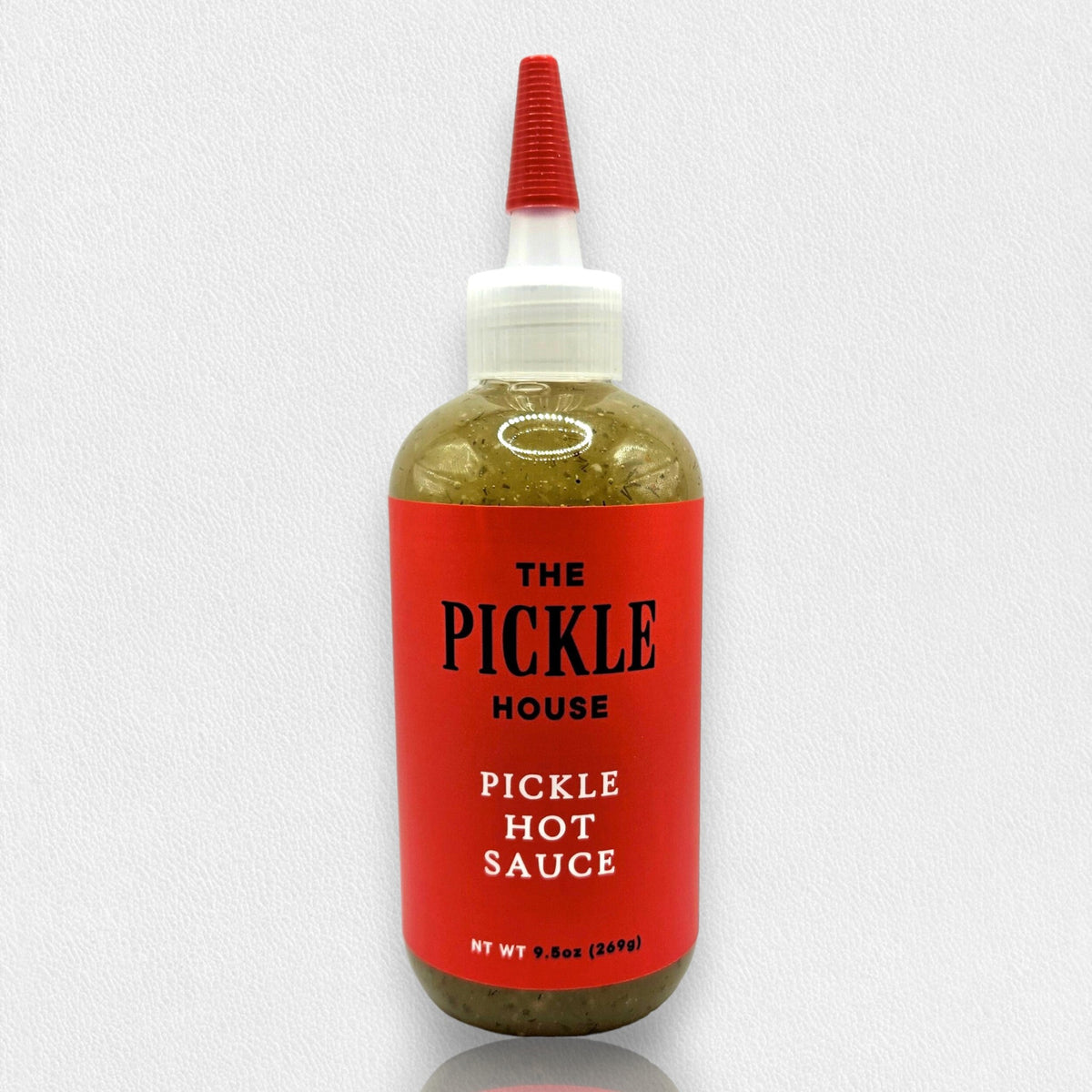 The product labeled "PICKLE HOUSE - PICKLE HOT SAUCE" from THE PICKLE HOUSE is displayed against a plain white background. The bottle, adorned with a red label featuring black text and capped with a white nozzle, contains 9.8oz (280g) of greenish-hued sauce. Its distinctive flavor arises from a secret spice blend mixed with tangy pickle brine.