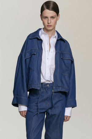 A person in a loose-fitting NUDE - DENIM JACKET by NUDE and matching jeans is seen from behind, standing in a shaded outdoor area.