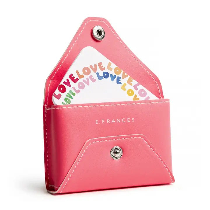 A person in a cream sweater holds the E. FRANCES - E. FRANNIE PACK, BUBBLEGUM wallet by E. FRANCES PAPER, pulling out a white card with red hearts—an adorable gift for spreading joy.
