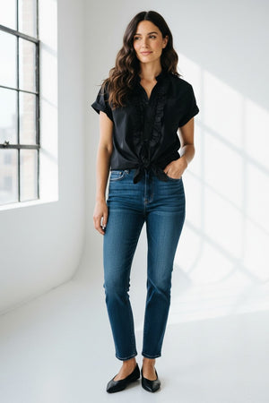 A woman wears the KMJ KIKI SHIRT, a black stand collar blouse with front ruffles, tucked into high-waisted blue jeans. She accessorizes with layered gold jewelry and red lipstick, hands resting on her hips. Brand: KMJ.