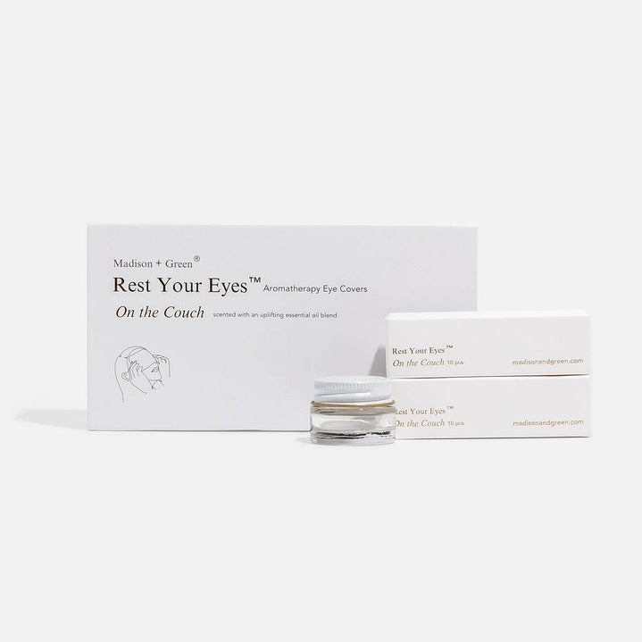 A person in a gray sweater holds an open white box containing MADISON GREEN’s “REST YOUR EYES ON THE COUCH,” lifting the small jar with a white and gold lid from the box—perhaps an essential oil blend designed for calming eye masks or aromatherapy eye covers.